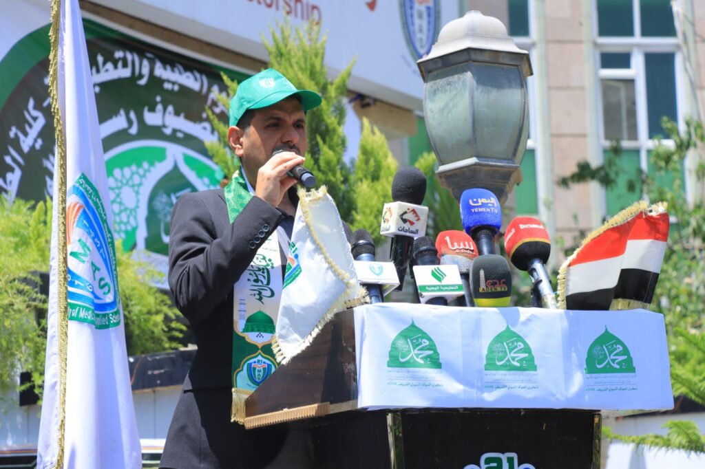 A man in a suit speaks into a microphone at a podium, surrounded by multiple news microphones and flags outdoors.