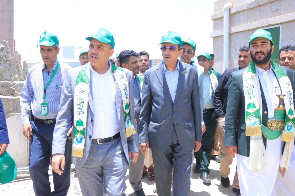 Group of men in suits and teal hats walk together outdoors, wearing ceremonial green sashes with logos beside a stone building.