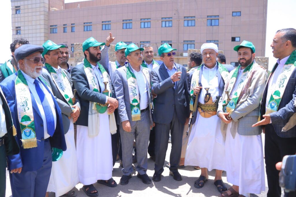 Group of men in traditional robes and green sashes posing together outdoors at a ceremony near a tan building.