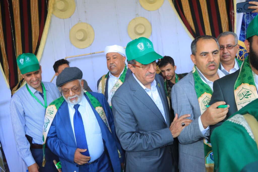 Group of men in suits and traditional attire wearing green caps and ceremonial sashes at a formal gathering, with decorative backdrops behind them.