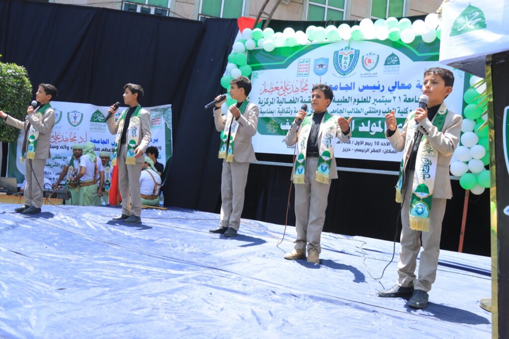 Group of five boys in beige suits singing with wired microphones on a blue stage, wearing green scarves at an outdoor event with Arabic banners and green balloons in the background.