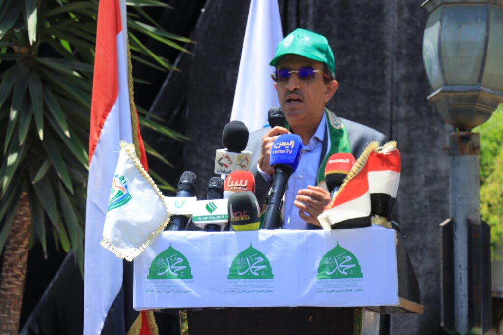 Man wearing a green cap and sunglasses speaks at a podium with multiple microphones and flags outdoors, delivering a speech.