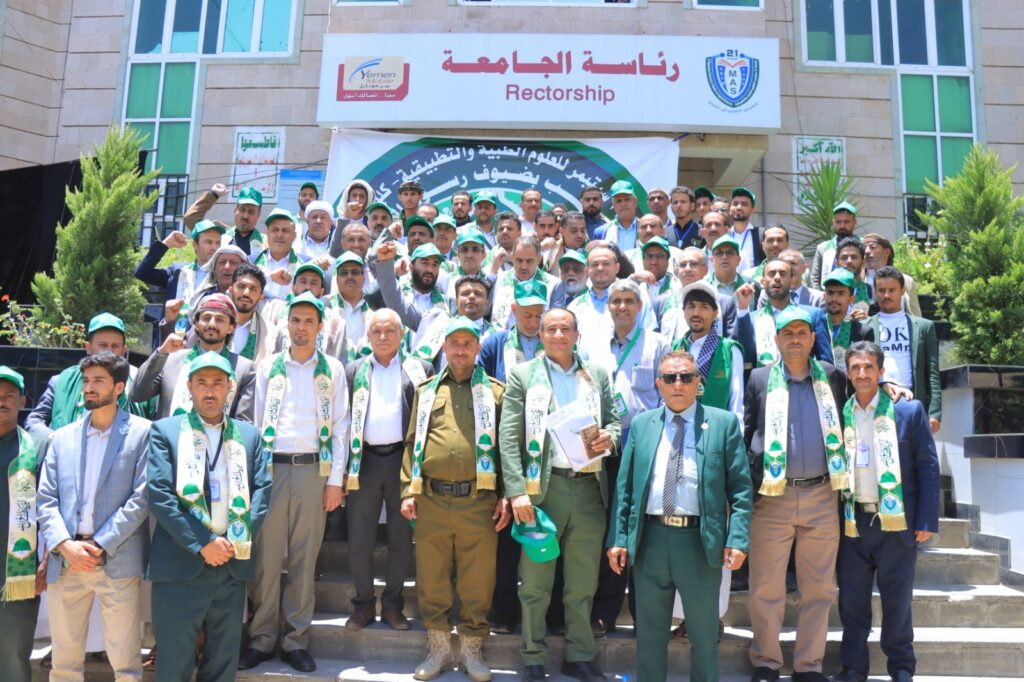 Group of university staff and students posing on stairs in front of a rectorate building, many wearing green scarves and caps; banner above reads Rectorship/رئاسة الجامعه.