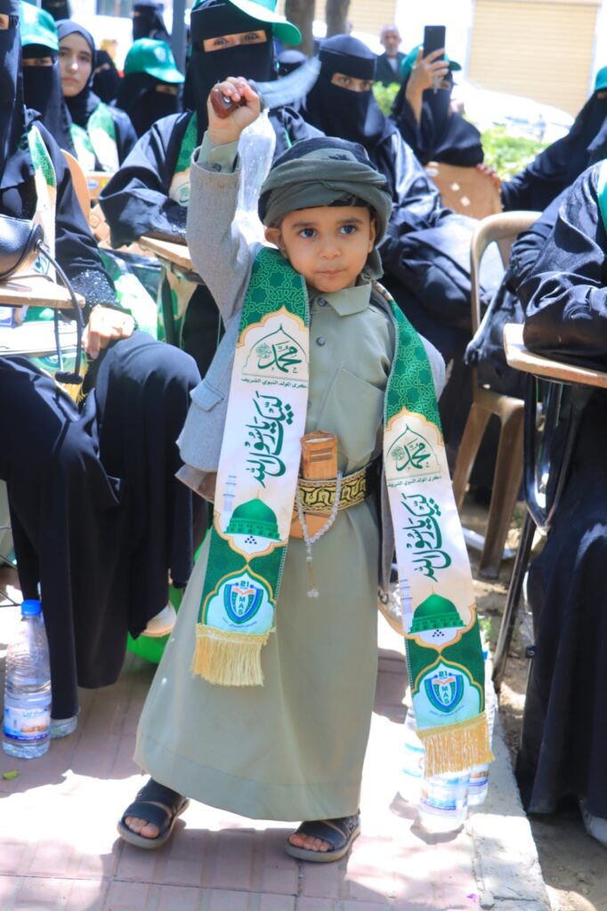 Young boy in green traditional clothing and head wrap raises his fist, wearing decorative sashes at an outdoor event with women in black abayas in the background.