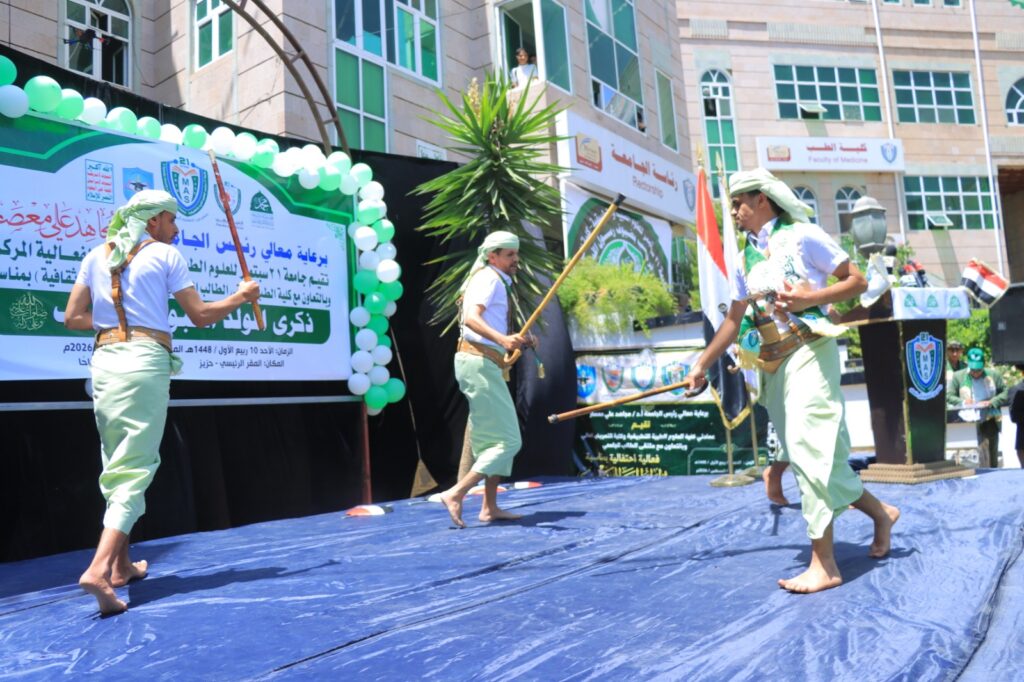 Three men in white shirts and light green pants perform a traditional sword dance barefoot on a blue stage, with a green-and-white banner and balloons behind them.