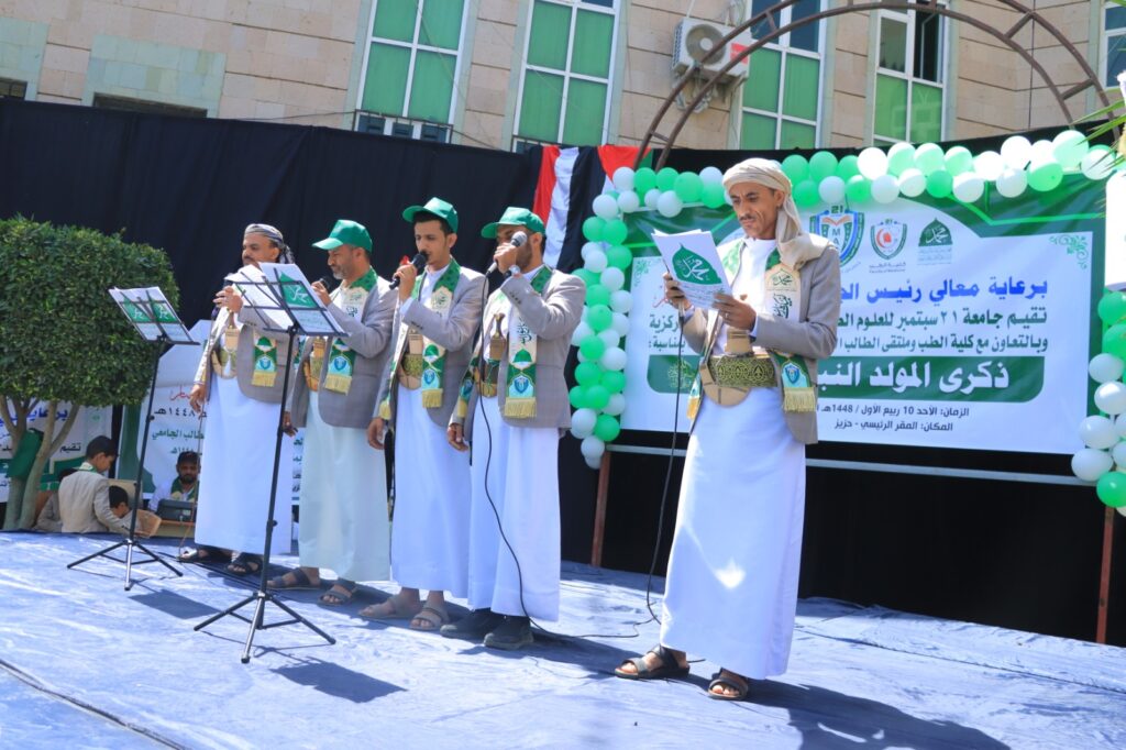 A group of men in traditional robes sings on a stage with microphones, green hats, and matching sashes, next to a banner and green balloons.