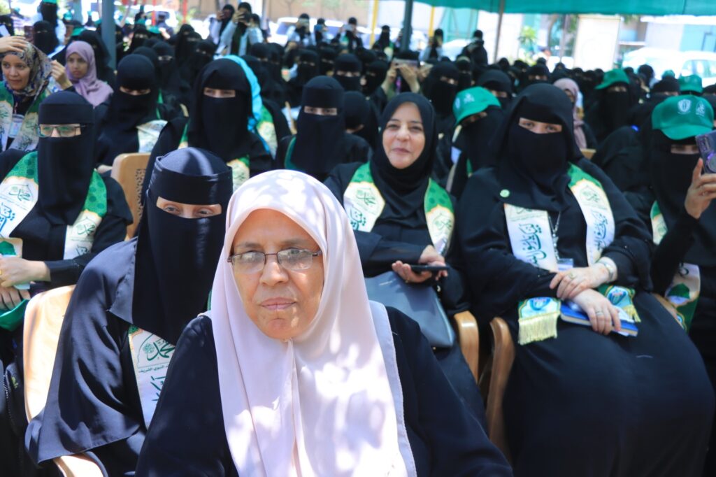 Front row woman in light purple hijab and glasses sits among a large crowd of women wearing black niqabs and green ceremonial sashes at an outdoor event.