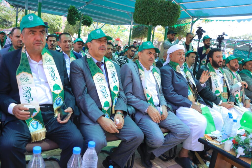 Group of men in suits wearing green ceremonial sashes and hats, seated at an outdoor event with cameras and other attendees in the background.