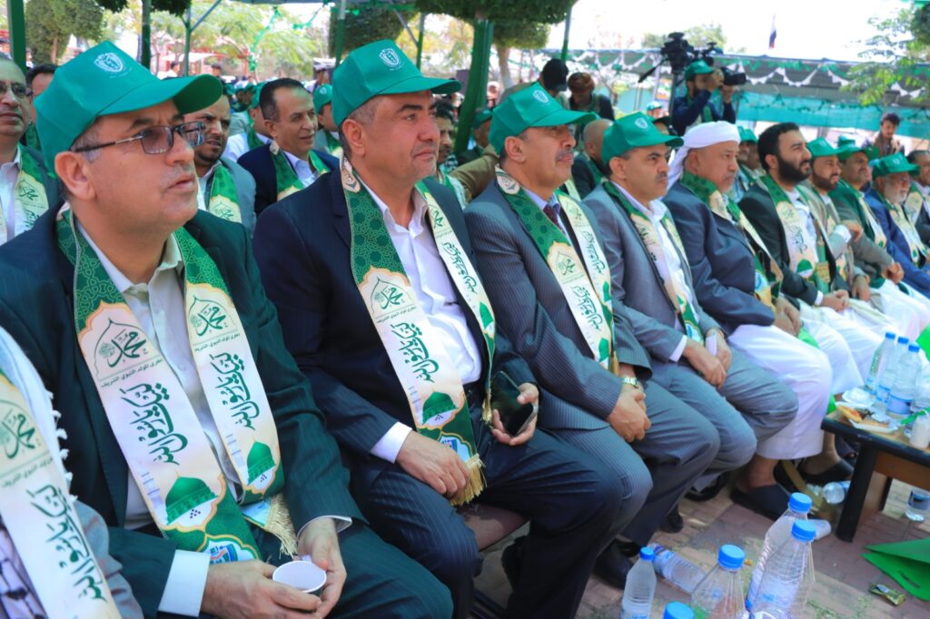 Group of men in suits with green ceremonial sashes and caps seated at an outdoor event, camera crew visible in the background.
