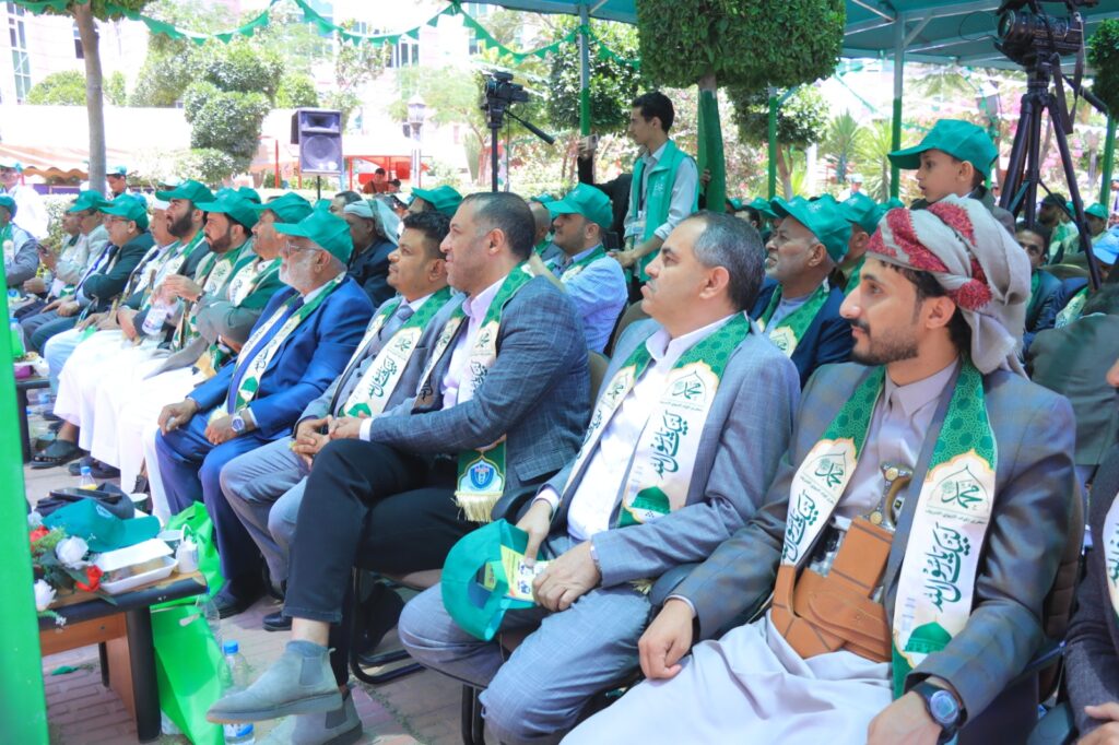 Group of men in suits and traditional attire seated in a row at an outdoor ceremony, wearing green scarves and hats under a shaded tent.