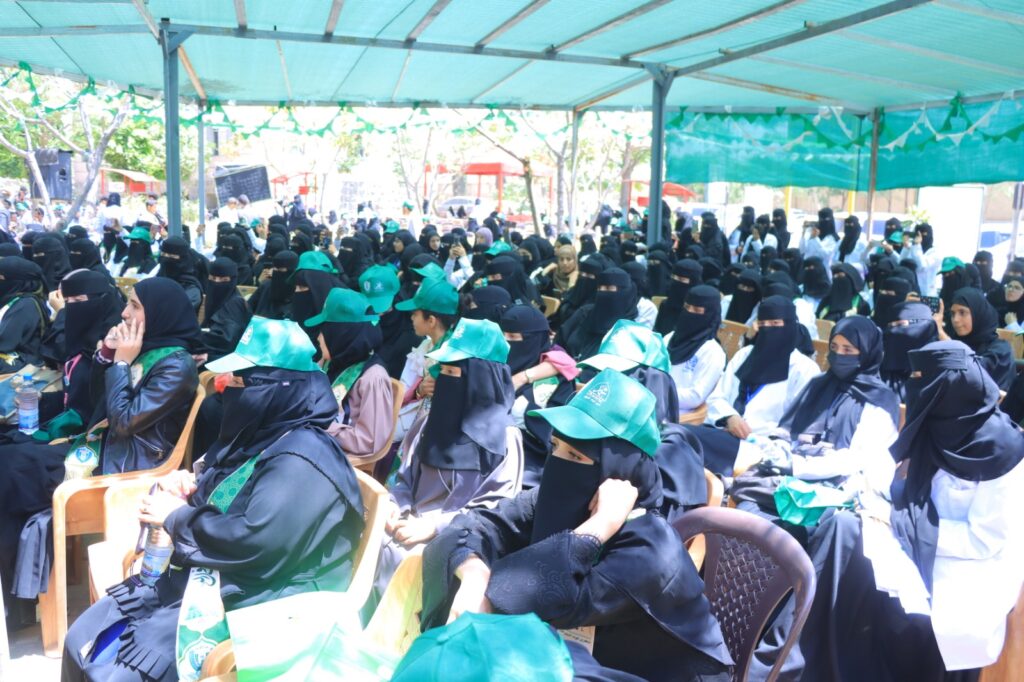 Large outdoor gathering of women wearing black niqab and green hats under a teal canopy, attending an event.