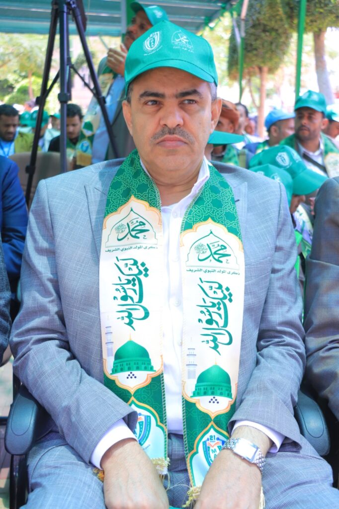 Man in a gray suit seated at an outdoor ceremony, wearing a green ceremonial scarf and cap with Arabic logos.