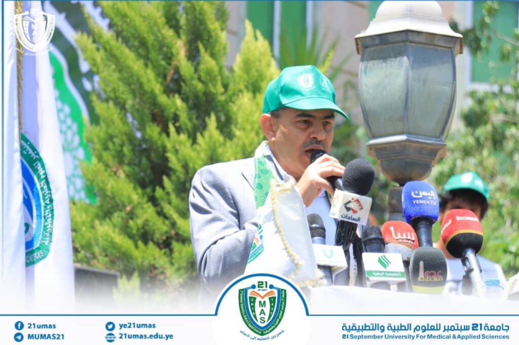 Man in a gray suit and green cap speaks into a microphone at an outdoor university event, with multiple media microphones in front and campus banners in view.