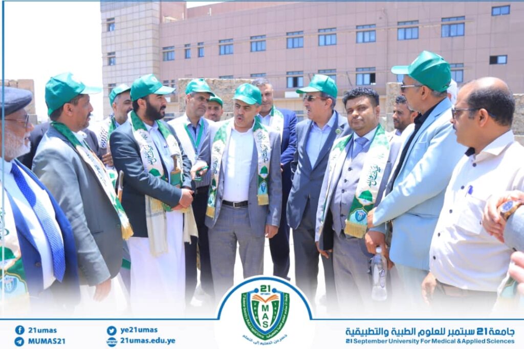 Group of men in green caps and scarves standing in a circle during a university event outdoors, blue banner with logo in front.