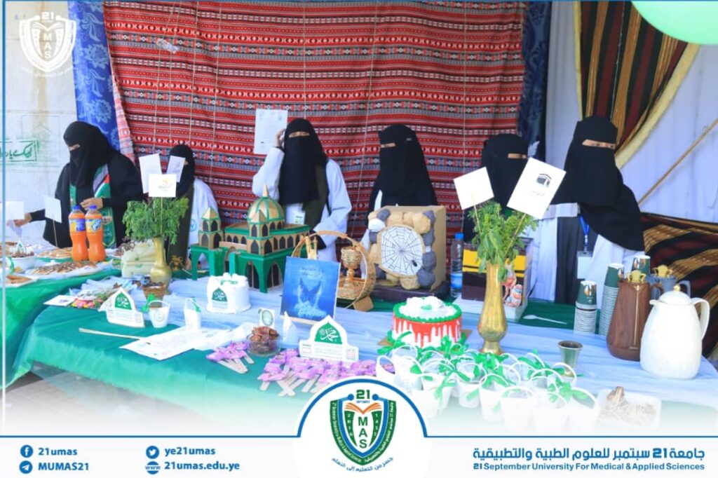 Group of women wearing niqab stand behind a long display table filled with models, plants, and foods at a university fair, with a patterned backdrop and banners showing the 21ums logo.