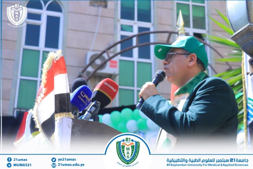 Man in a green cap and suit speaks into a microphone at a podium during a university event, with multiple media microphones in front and a campus backdrop.