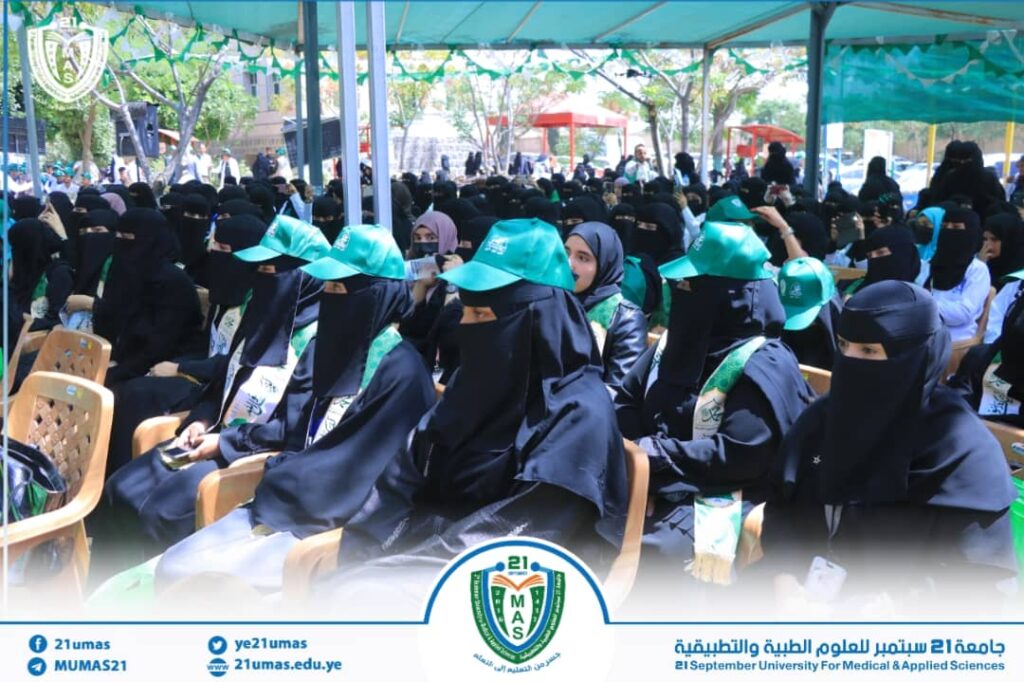 Group of women in black abayas and niqabs seated under a canopy at an outdoor university event; some wear turquoise caps.
