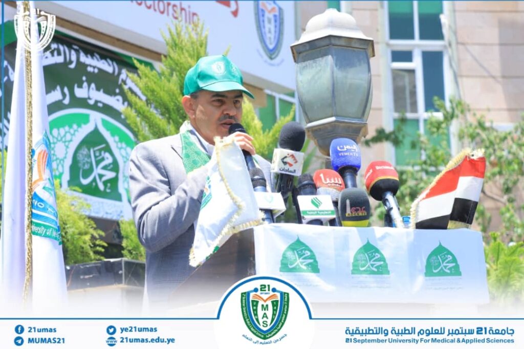 Man in a gray suit and green cap speaks into multiple microphones at an outdoor university event.