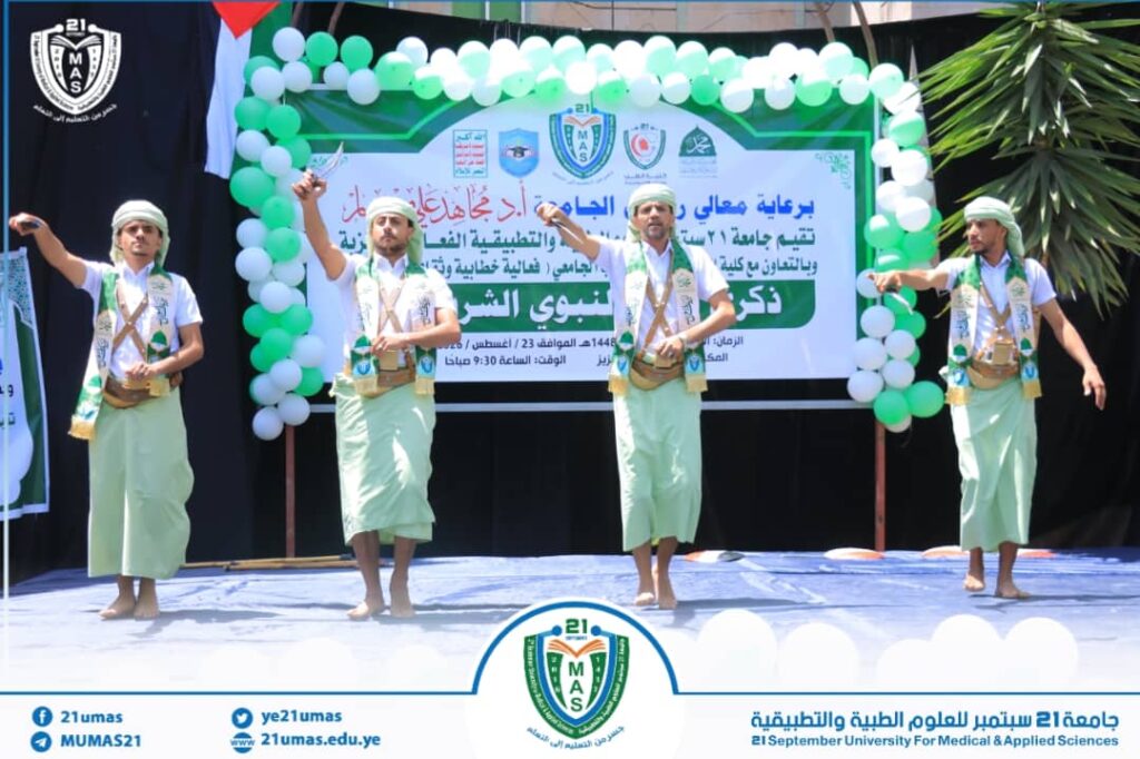 Four male dancers in traditional green outfits perform on a decorated stage with a green‑white balloon arch and a large banner behind them at a university event under a sunny outdoor setting.