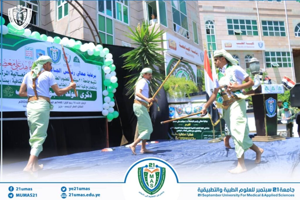 Three dancers in traditional Yemeni attire perform a sword dance on an outdoor university stage with banners and the campus crest in the background.