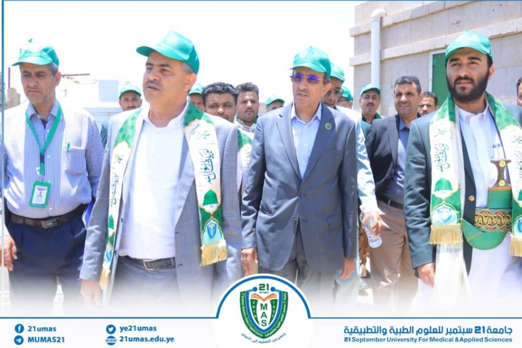 Group of men in suits and green caps outdoors at a ceremony, wearing banners and sashes with a university emblem visible at the bottom.
