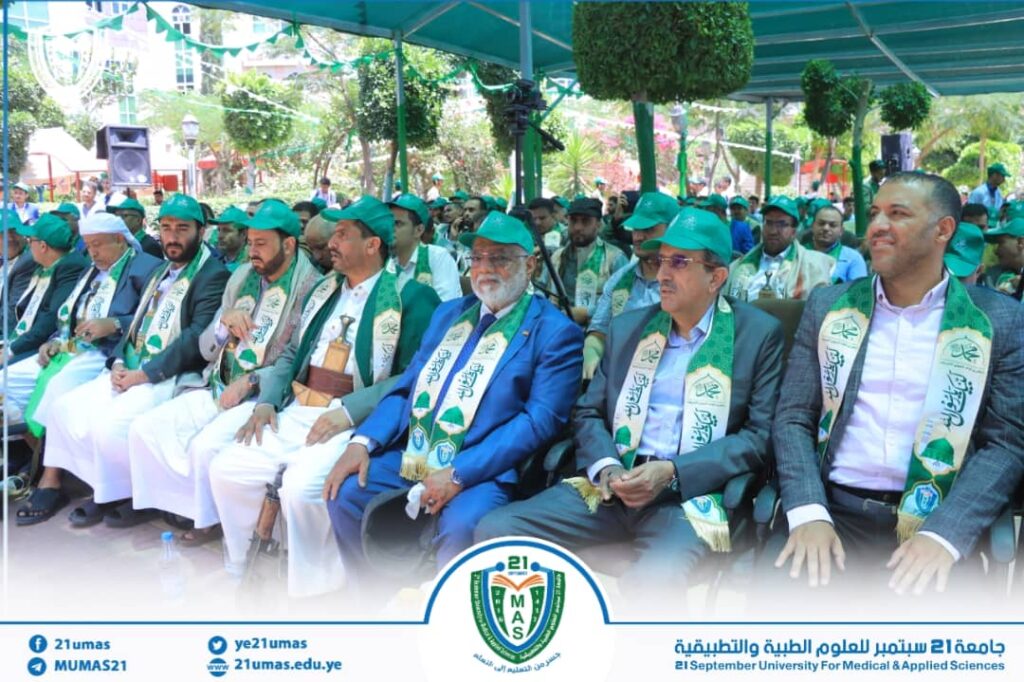 Group of men in green caps and matching scarves seated outdoors at a university ceremony, with a banner and university logo visible at the front.