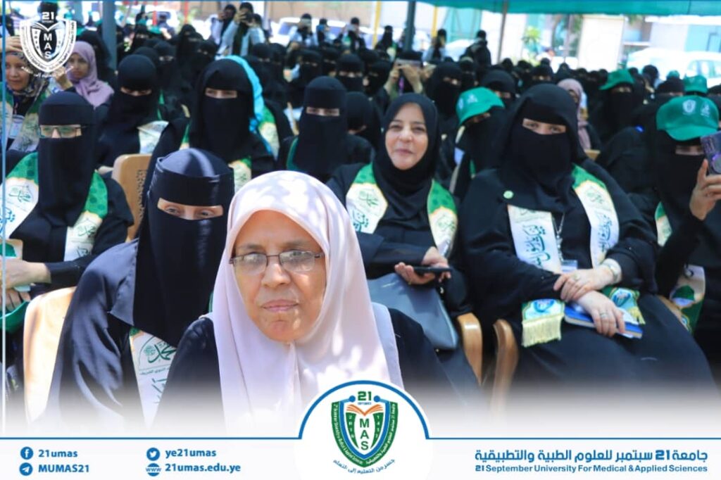 Group of women in black abayas at an outdoor ceremony, many wearing green sashes; foreground woman in a light hijab and glasses.