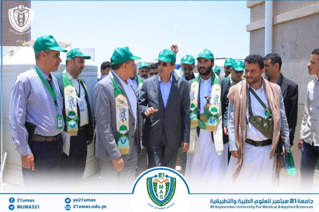 Group of men in suits and green caps with ceremonial sashes at an outdoor university event, logo of 21 September University for Medical & Applied Sciences in view.