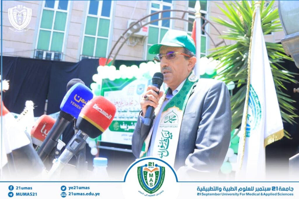 Man in a suit and green cap speaks into a handheld microphone at a university event with banners and microphones in front of him.