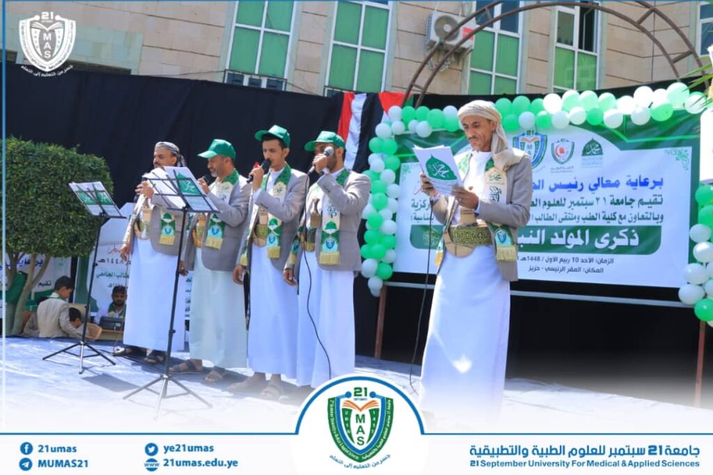 Six men in traditional attire sing into microphones on a university stage with green-white balloons and a banner in the background.