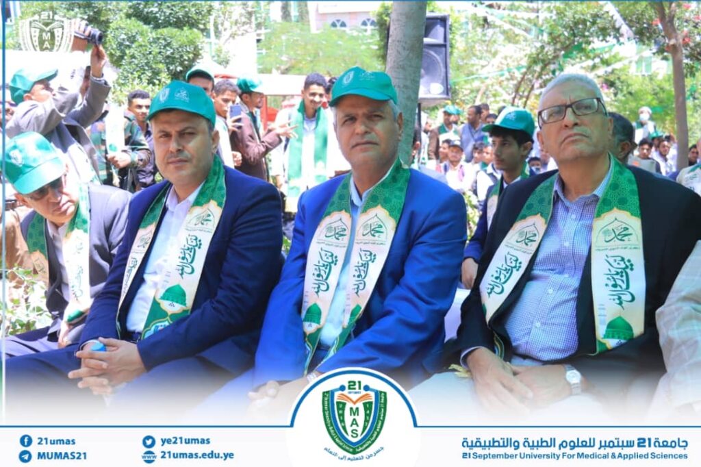 Four men in blue suits with green scarves and matching caps sit at an outdoor university ceremony, with 21 September University branding visible in the background/rear banners.