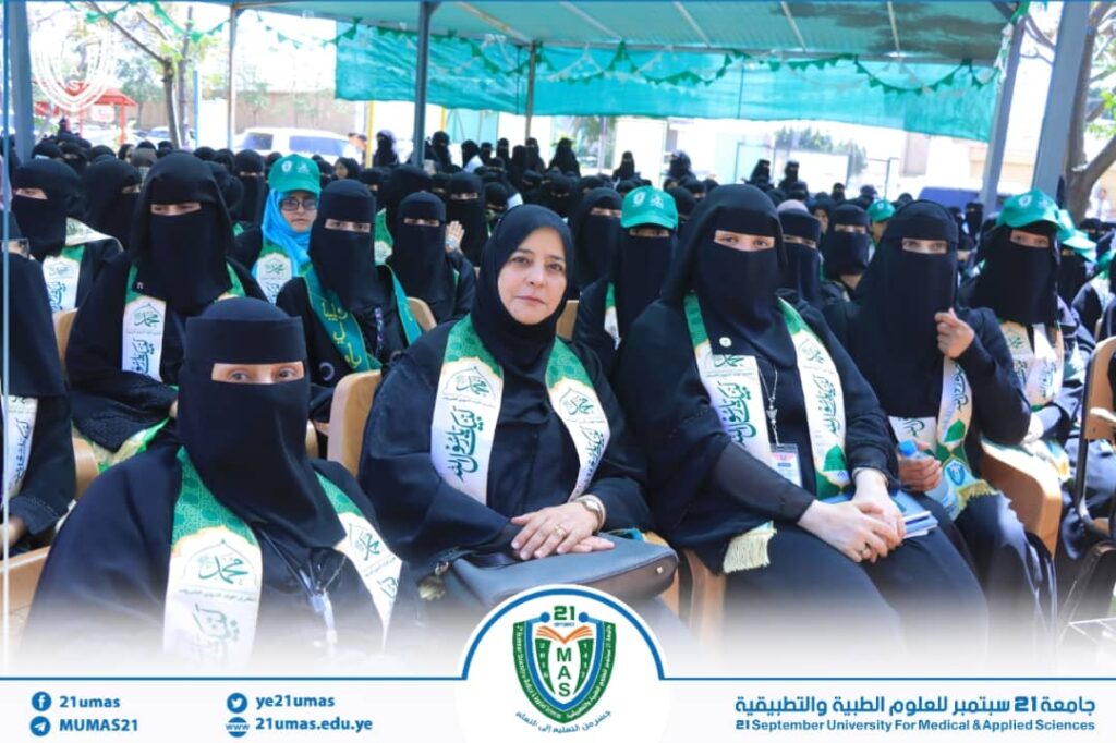 Group of women in black abayas and niqabs seated at an outdoor ceremony under a turquoise canopy, wearing green and white sashes; university crest visible at the bottom indicating 21 September University for Medical & Applied Sciences.