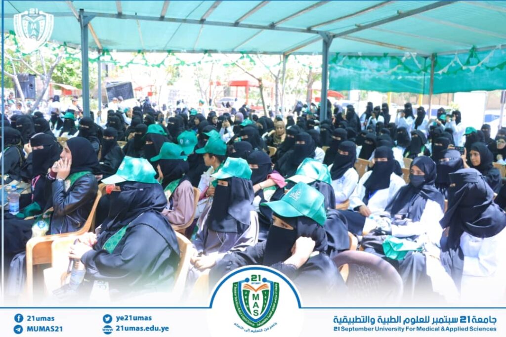 Large group of women in black hijabs and green caps seated under a blue canopy at a university event.