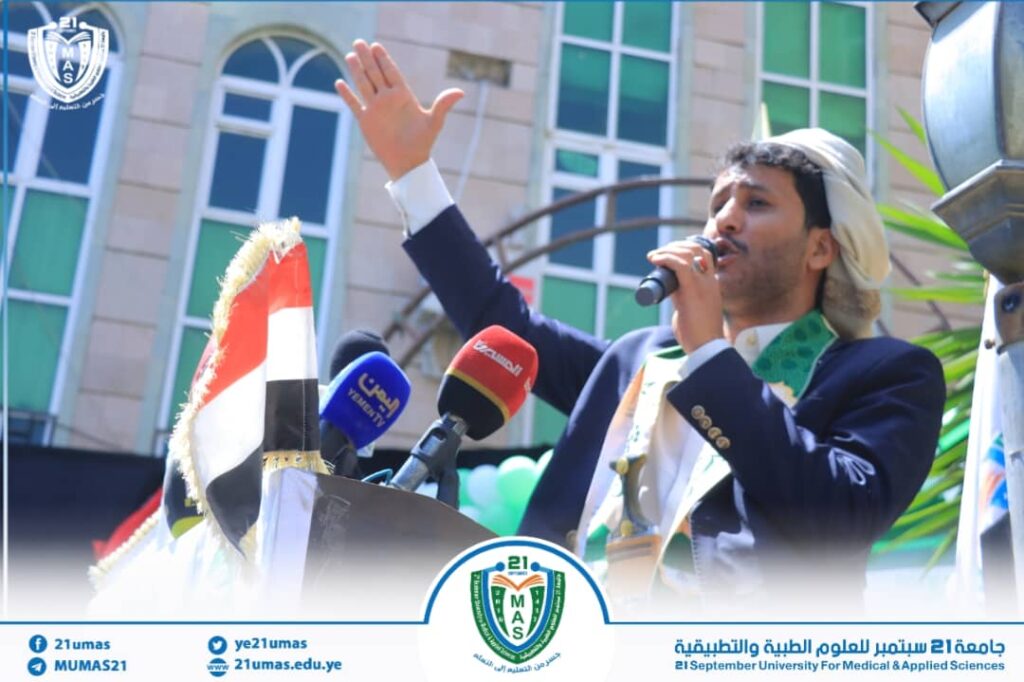 Man in a suit speaks into a microphone at a podium, raising his left hand, with the Yemeni flag and university logo nearby (outdoor event).