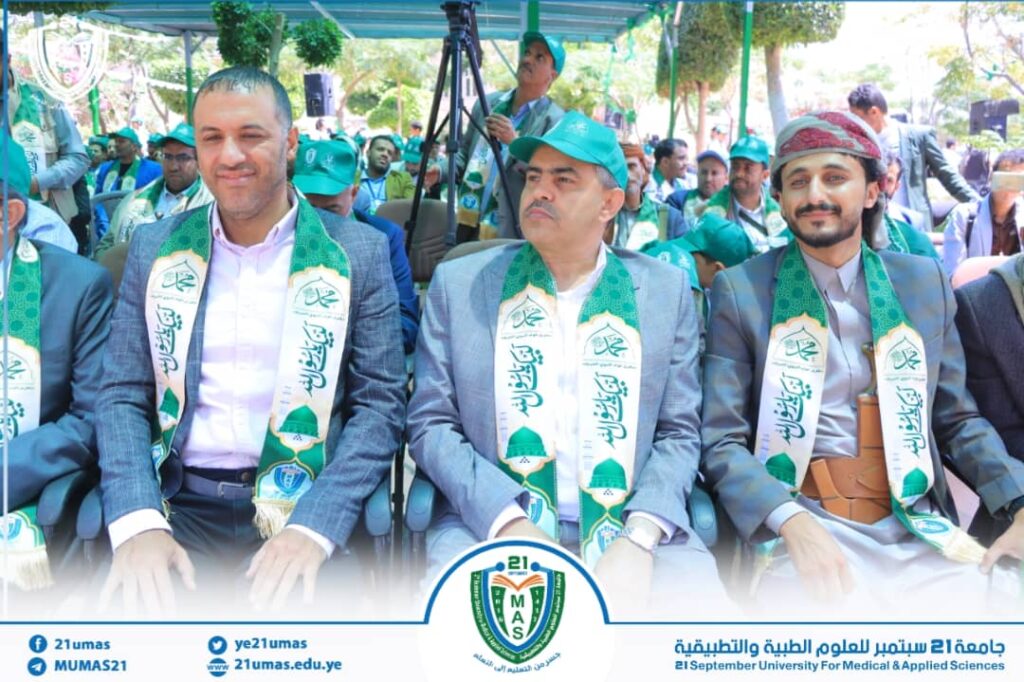Three men in suits wearing green-and-white ceremonial sashes sit in a row at an outdoor university event, with a crowd and a banner visible in the background