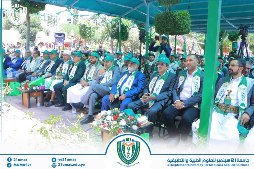 Group of formally dressed attendees seated outdoors under a teal canopy, wearing green scarves with a university emblem for a campus event.