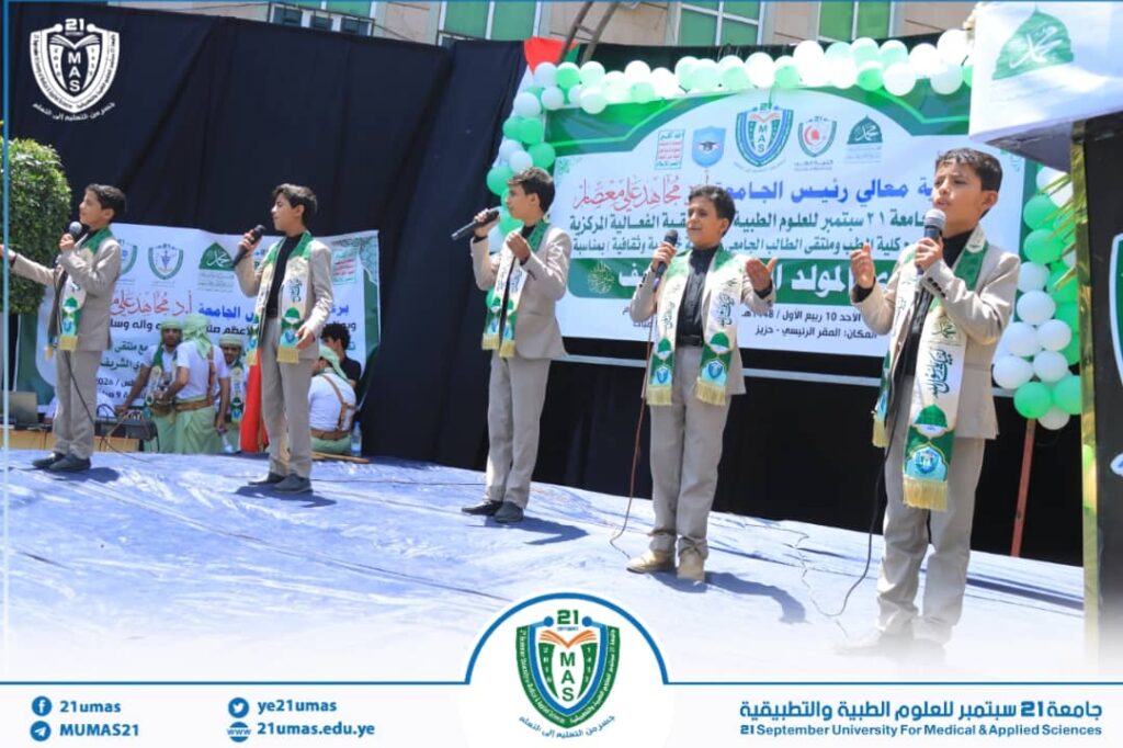 Six young students in beige suits sing into microphones on an outdoor stage during a university event, with a green-and-white banner and balloons in the background.