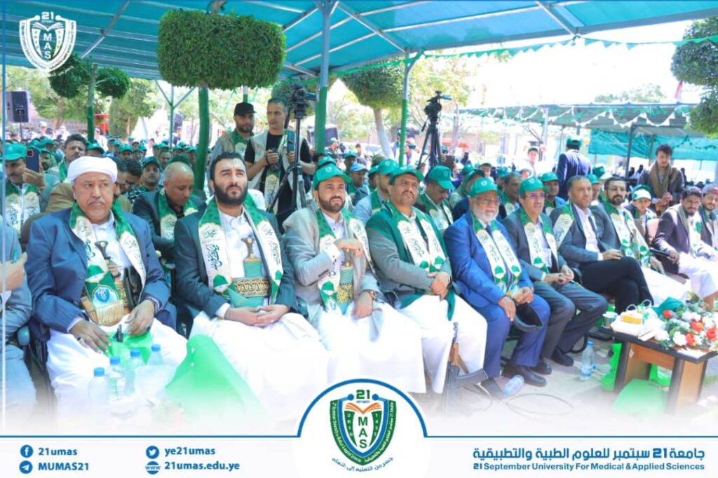 Group of men in suits with green scarves seated at an outdoor ceremony under blue canopies, with a university banner in view.