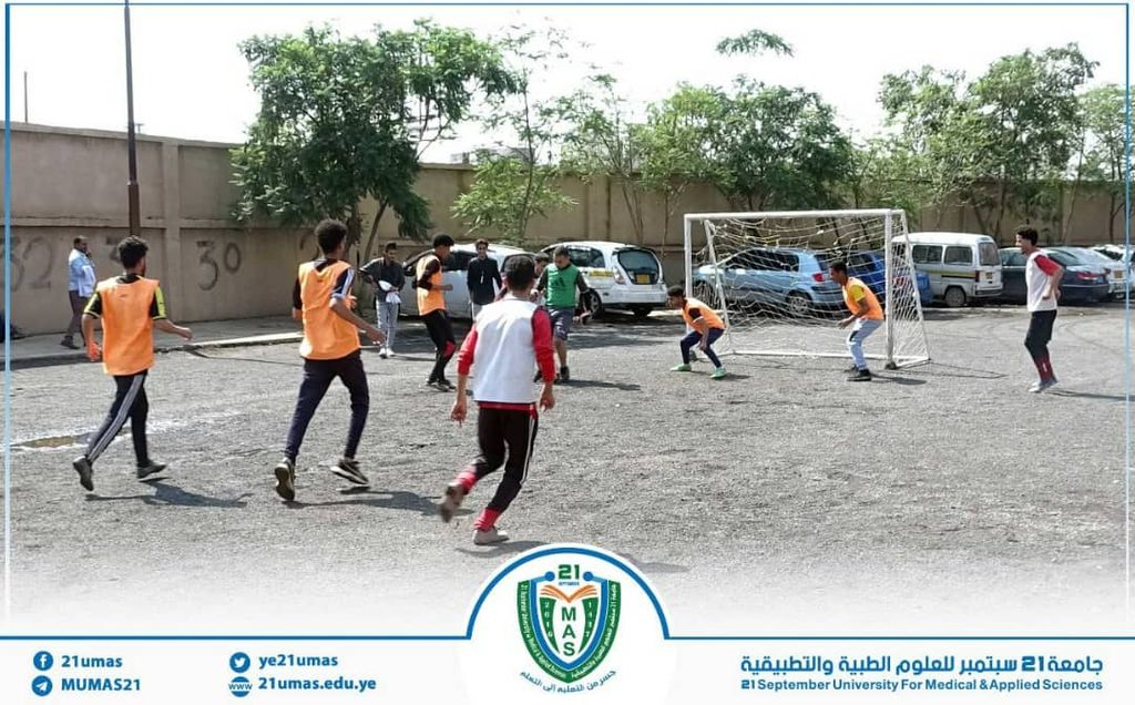 Group of students in orange pinnies playing soccer on a gravel lot with a metal goal; campus cars and trees in the background, university banner at bottom.