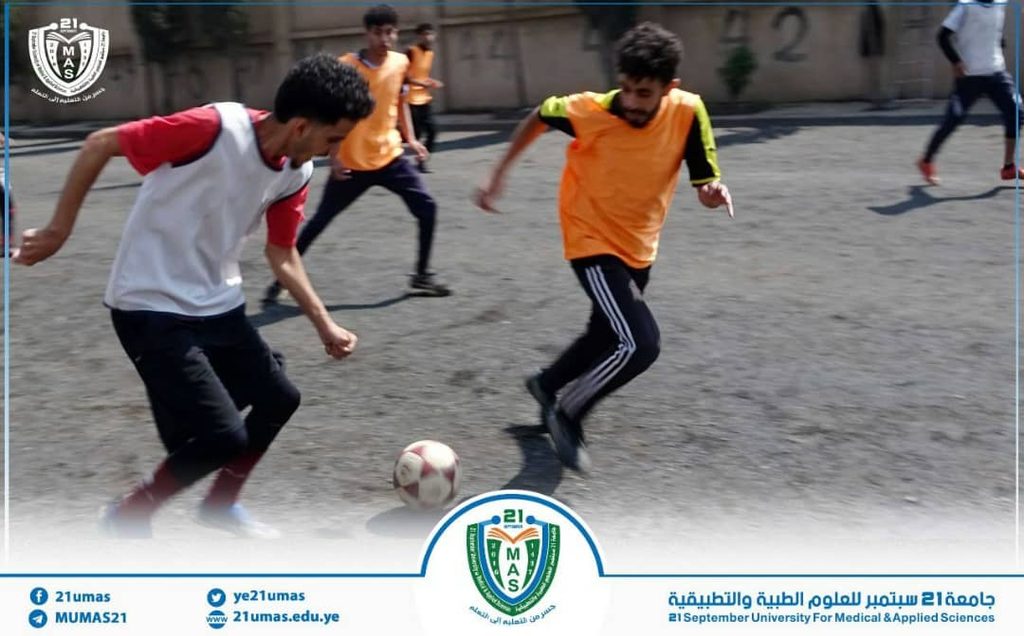 Young men playing soccer on an outdoor court, with a ball near the center; university logo and branding at the bottom.