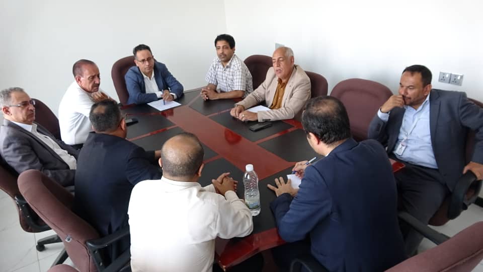Eight men in business attire sit around a rectangular conference table in a bright office, engaged in a meeting with notes and a water bottle at the center.