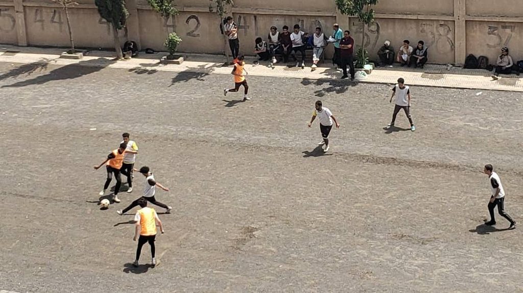 IMG-20260830-WA0135 - جامعة 21 سبتمبر Young players in orange bibs and white shirts play soccer on a dusty outdoor court, while spectators sit along a graffiti-covered wall nearby.