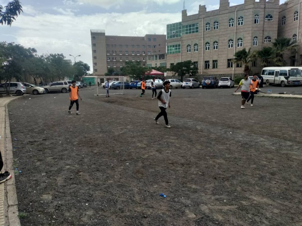 IMG-20260830-WA0136 - جامعة 21 سبتمبر Group of people playing soccer on a gravel field near a campus building and parked cars.