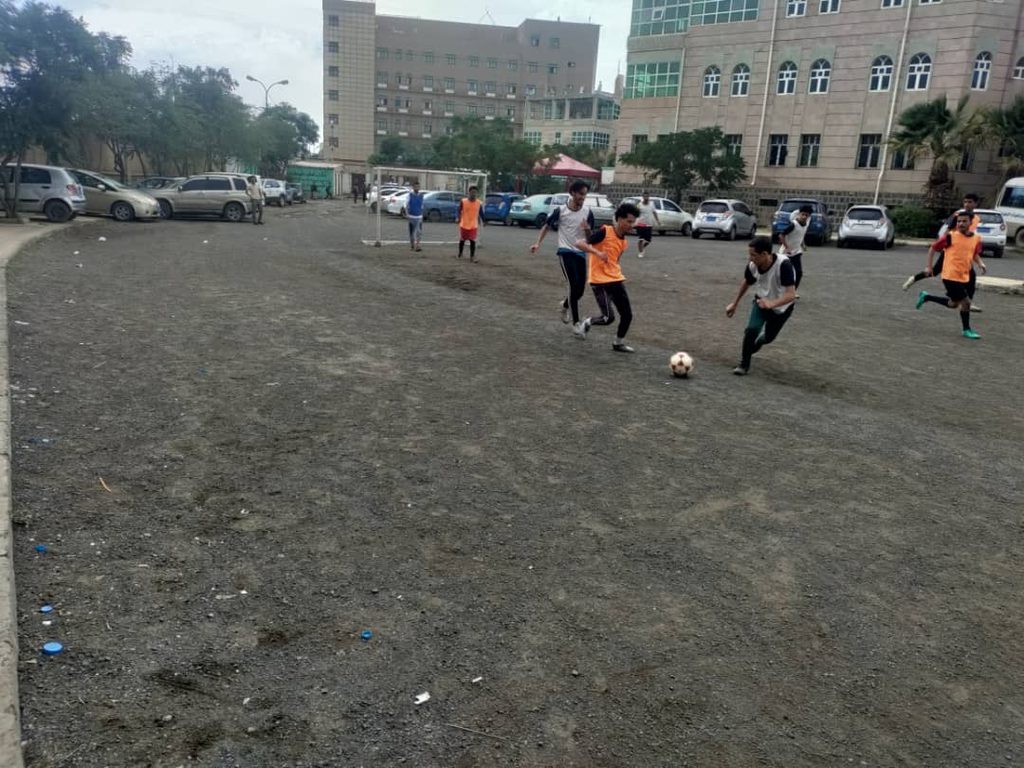 IMG-20260830-WA0138 - جامعة 21 سبتمبر Group of kids playing soccer on a gravel field, with parked cars and campus buildings in the background.