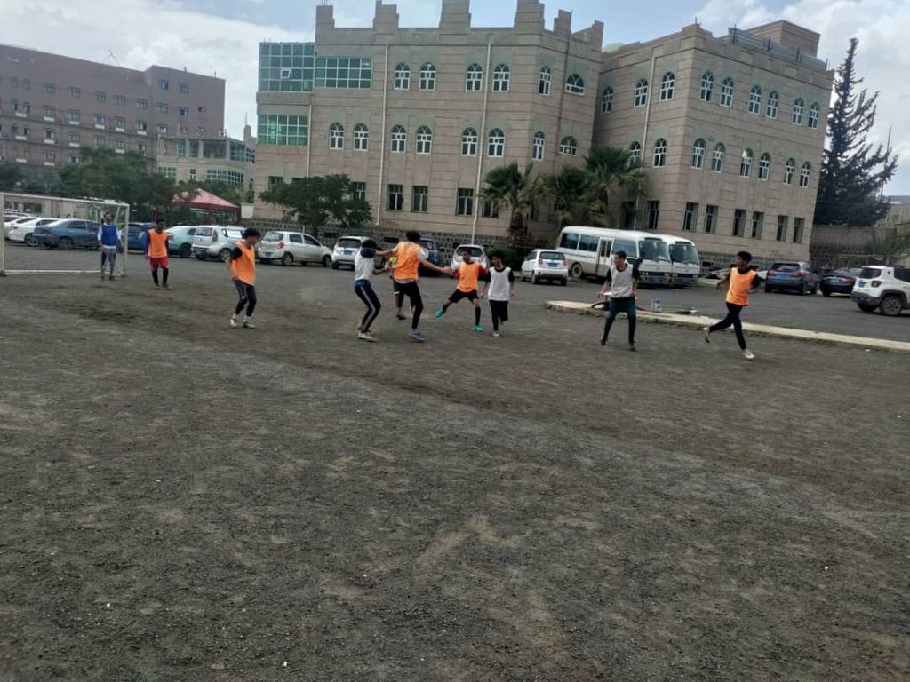 IMG-20260830-WA0139 - جامعة 21 سبتمبر Group of people playing soccer on a dirt field in front of a large beige building, some wearing orange pinnies.