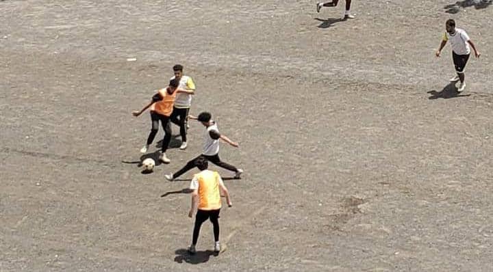 IMG-20260830-WA0140 - جامعة 21 سبتمبر Group of young people playing soccer on a dusty, open field; one player in an orange bib challenges for the ball with others nearby and a teammate watches from behind.
