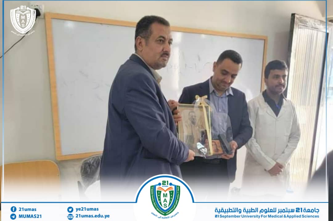 Two men in suits presenting a glass-framed award to another man, with a third person in a white coat watching nearby in a classroom setting.