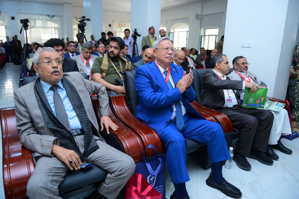 IMG_20260208_072653_657 - جامعة 21 سبتمبر A row of men in suits seated at a conference, with cameras and attendees in the background.