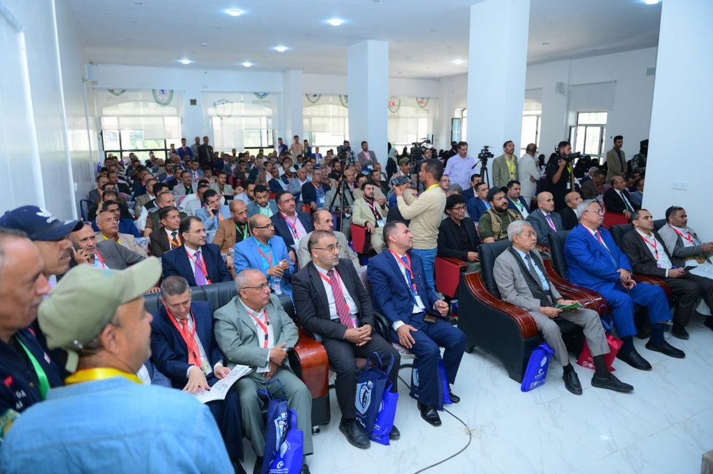 IMG_20260208_072653_833 - جامعة 21 سبتمبر Large conference hall full of attendees seated and listening, with photographers at the back capturing the event.