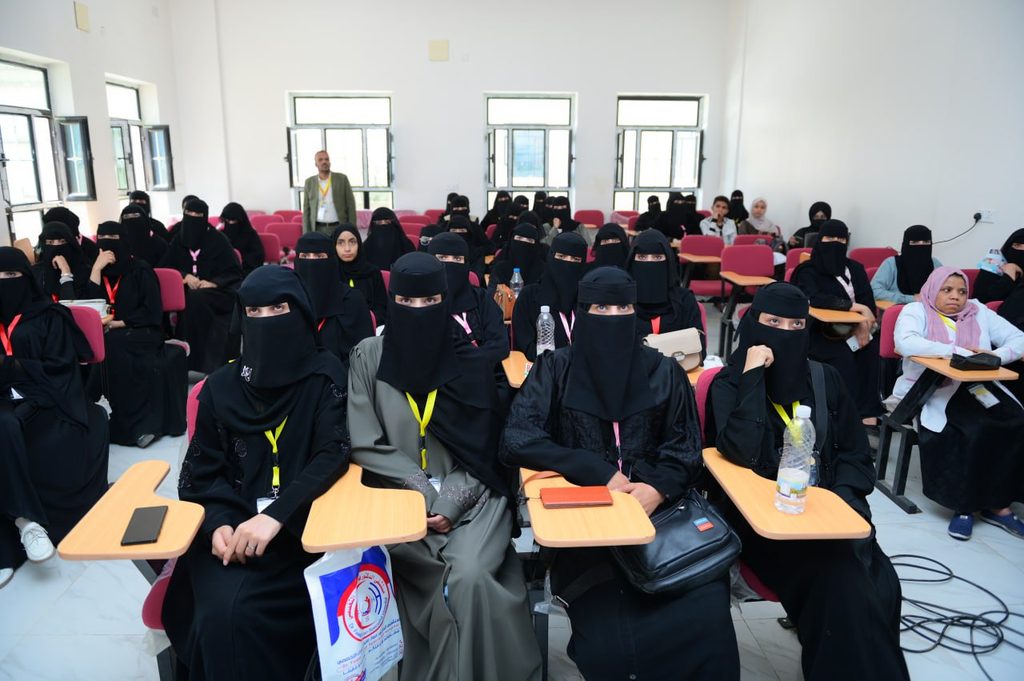 IMG_20260208_072702_208 - جامعة 21 سبتمبر Lecture hall filled with women in black abayas and niqabs seated at small desks; a man stands at the back of the room.